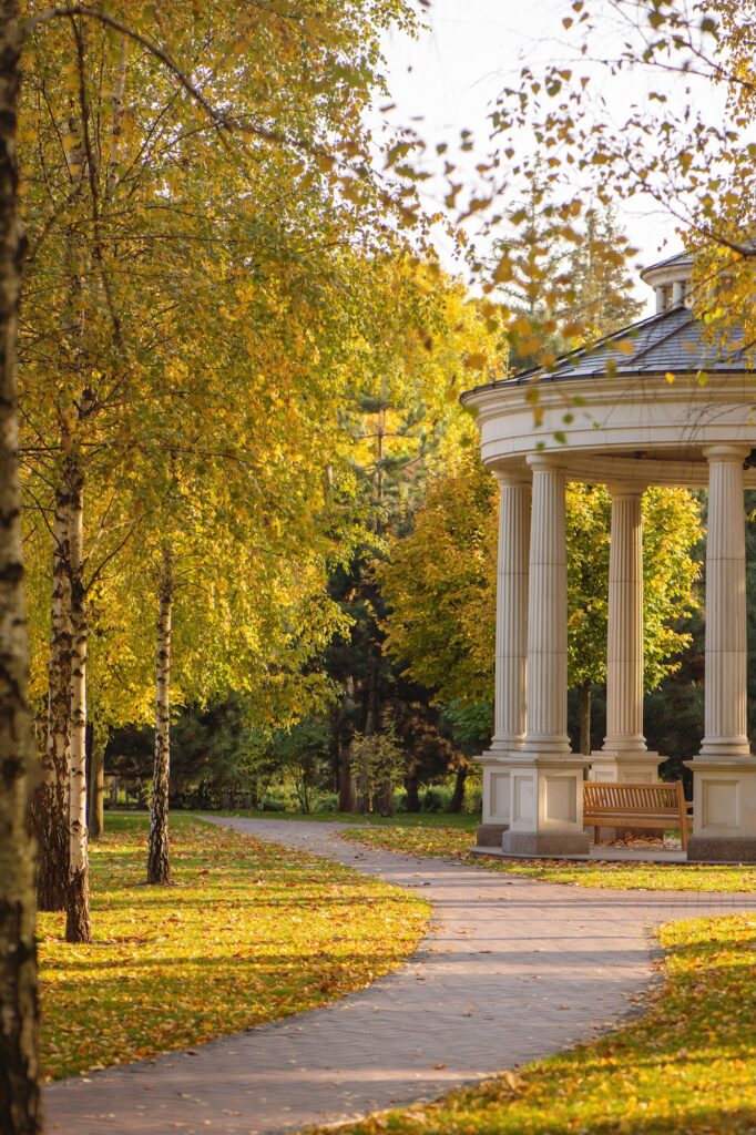 A paved path curves past a white, columned gazebo surrounded by trees adorned with vibrant Virginia fall foliage in shades of green and yellow. Sunlight gently filters through the autumn leaves, casting dappled patterns along the walkway.