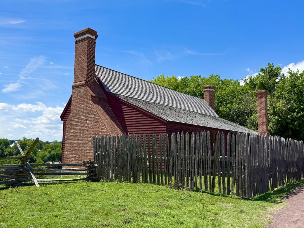 A red wooden house with two brick chimneys, reminiscent of classic presidential homes, is surrounded by a tall wooden fence and green grass under a bright blue sky.