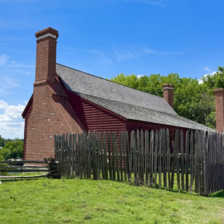 A red wooden house with two brick chimneys, reminiscent of classic presidential homes, is surrounded by a tall wooden fence and green grass under a bright blue sky.