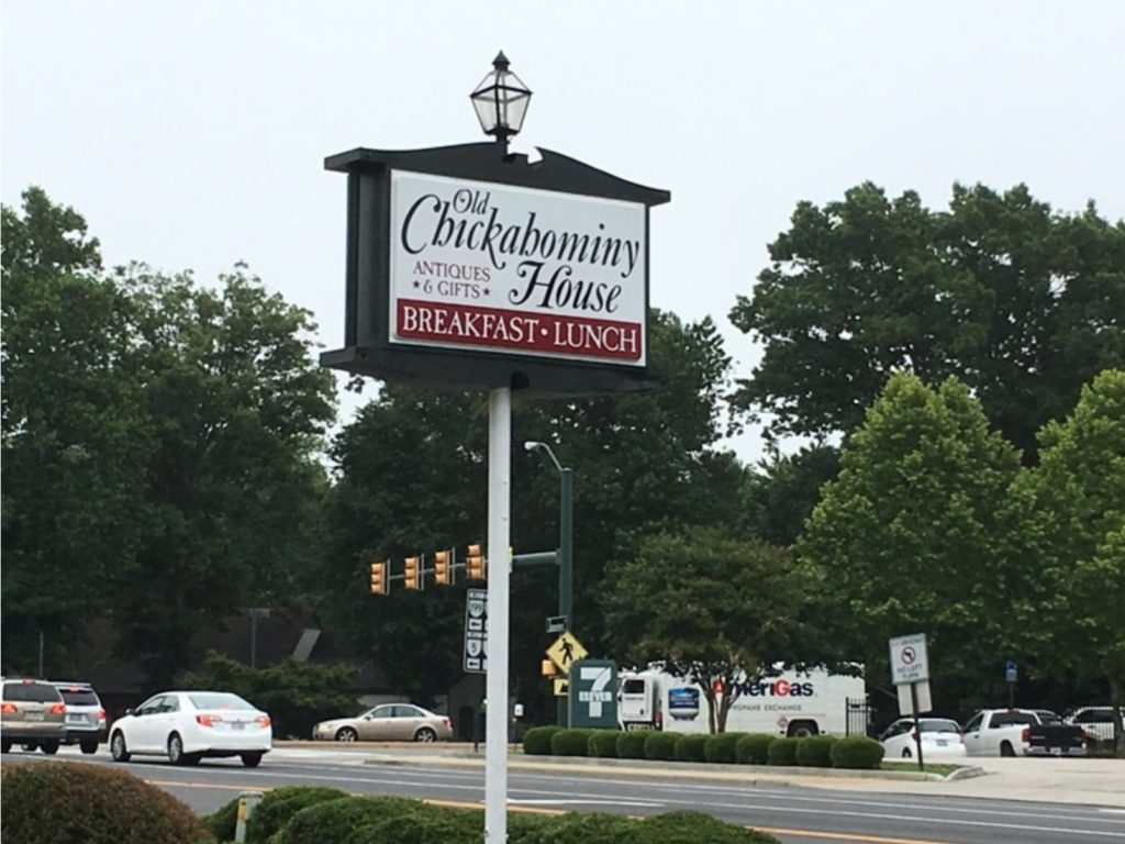 A sign for Old Chickahominy House, near historic Jamestown, advertises antiques, gifts, breakfast, and lunch. It stands by a busy street with trees and vehicles in the background.
