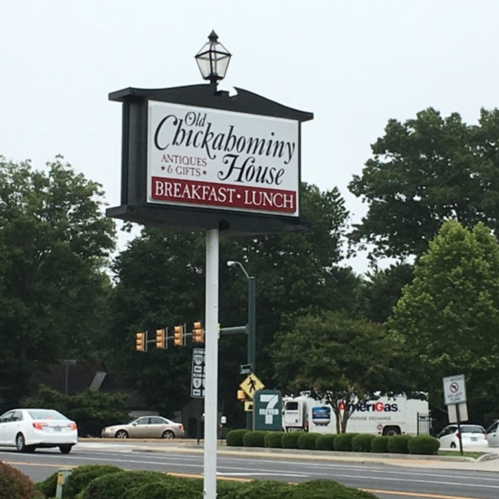A sign for Old Chickahominy House, near historic Jamestown, advertises antiques, gifts, breakfast, and lunch. It stands by a busy street with trees and vehicles in the background.