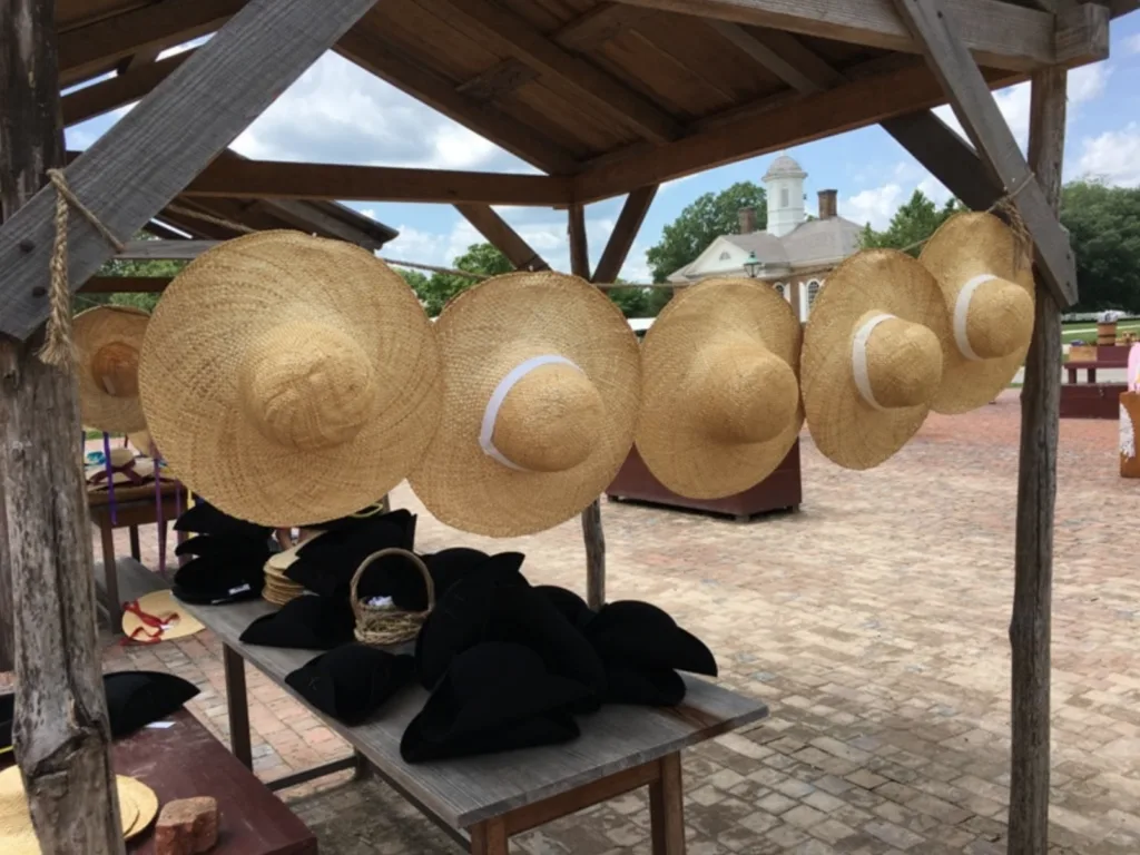 Large straw hats are hanging in a row under a wooden structure at an outdoor marketplace in Jamestown. Black tricorne hats are displayed on the table below. A white building is visible in the background.