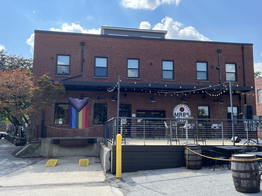 Exterior of a two-story red brick building with outdoor seating, string lights, and a pride flag on the patio railing. The building has a round sign that reads "MARS.