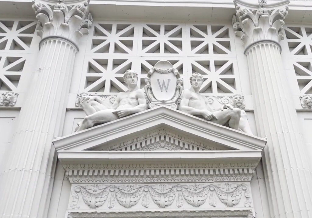 Two sculpted male figures sit beside a shield with a “W” on the neoclassical building facade in Front Royal, featuring columns, a decorative frieze, and geometric patterned relief.