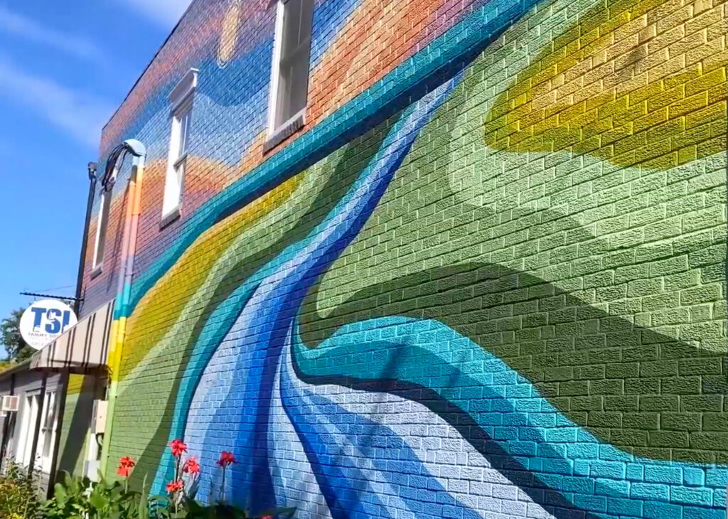 Colorful mural with flowing blue, green, and yellow shapes painted on a brick wall of a building in Front Royal, with red flowers in the foreground.