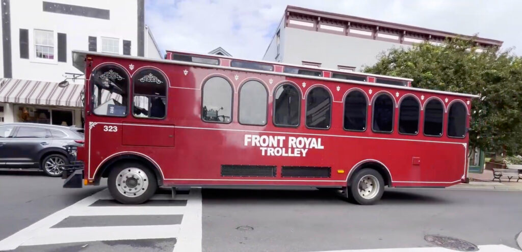 A red trolley labeled “Front Royal Trolley” is stopped at a crosswalk on a city street in the heart of Front Royal, with buildings visible in the background.