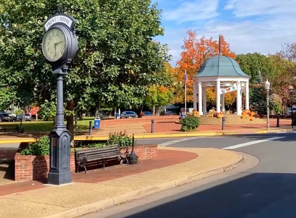 Town square in Front Royal with a vintage clock, bench, leafy trees, and a white gazebo underneath a blue sky. Orange autumn leaves are visible in the background.