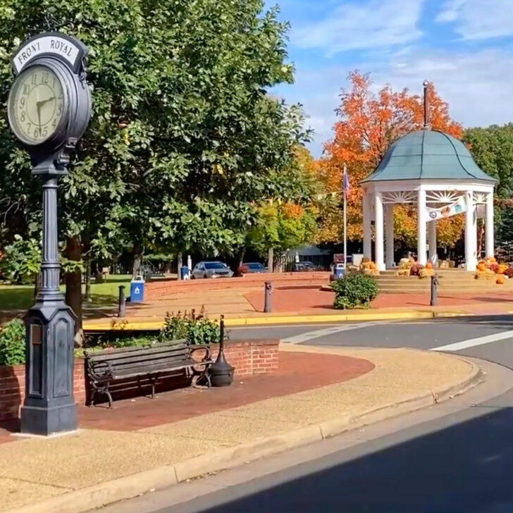 Town square in Front Royal with a vintage clock, bench, leafy trees, and a white gazebo underneath a blue sky. Orange autumn leaves are visible in the background.