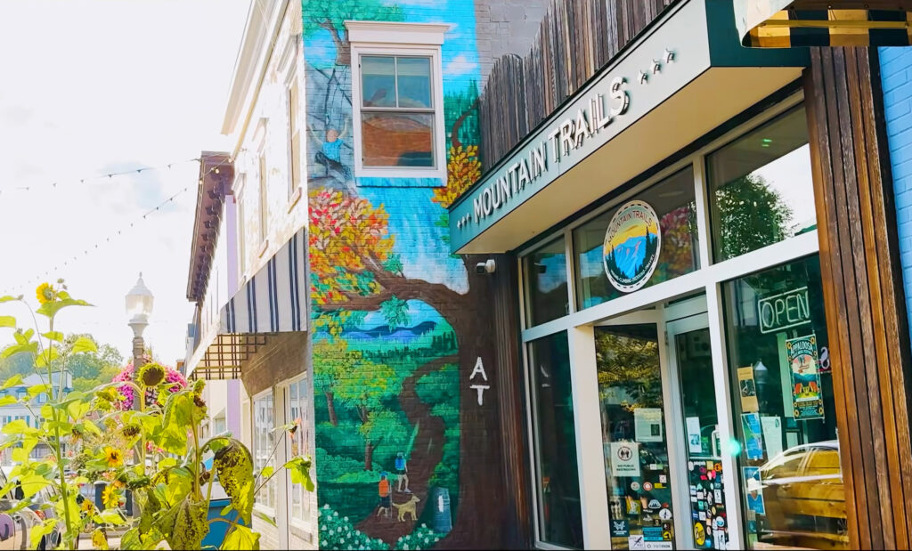 Outdoor mural of a tree and mountains on the side of a building next to the entrance of Mountain Trails store in Front Royal, with an “OPEN” sign on the glass door and a street scene reflected in the window.