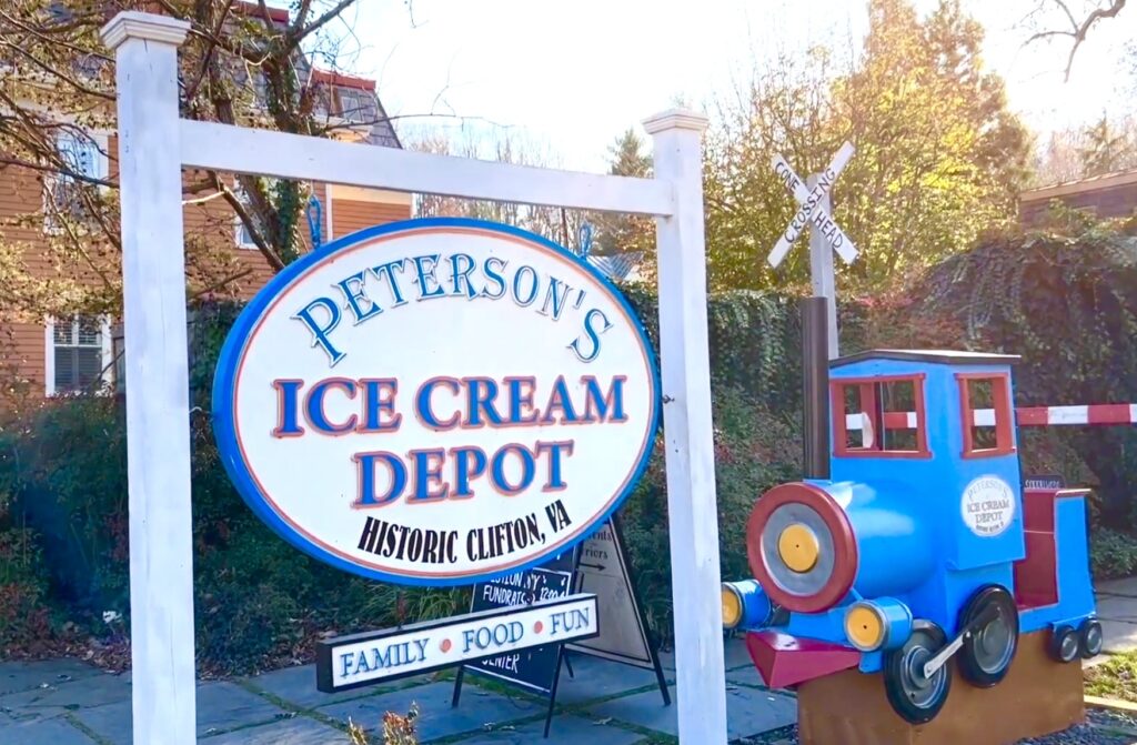 A sign for Peterson's Ice Cream Depot, a must-visit spot when searching for where to eat in Clifton VA, stands next to a blue and red toy train structure on a sunny day.