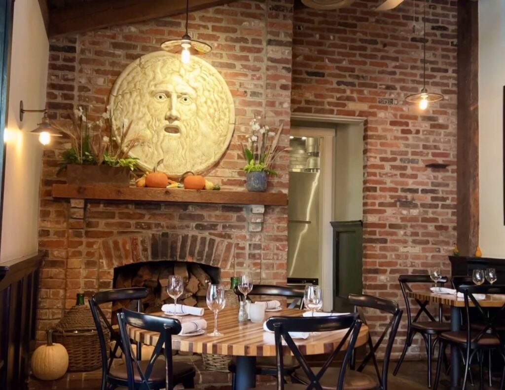 A cozy restaurant in Clifton VA featuring brick walls, set dining tables, and a fireplace adorned with plants, pumpkins, and a large circular face sculpture&mdash;perfect for those seeking where to eat in Clifton VA.