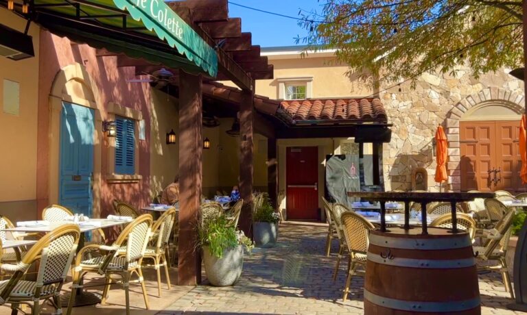 Outdoor café patio with wicker chairs and tables, shaded by a wooden pergola and surrounded by potted plants and stone buildings—a perfect spot if you're wondering where to eat in Clifton VA on a sunny day.