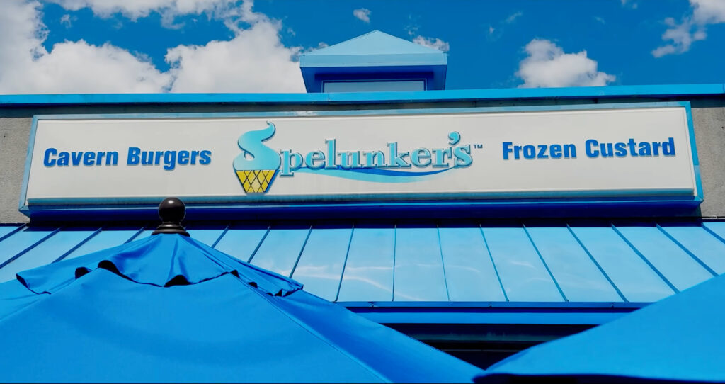 A restaurant sign reads “Cavern Burgers Spelunker’s Frozen Custard” above a blue roof and blue patio umbrellas in Front Royal, under a partly cloudy sky.