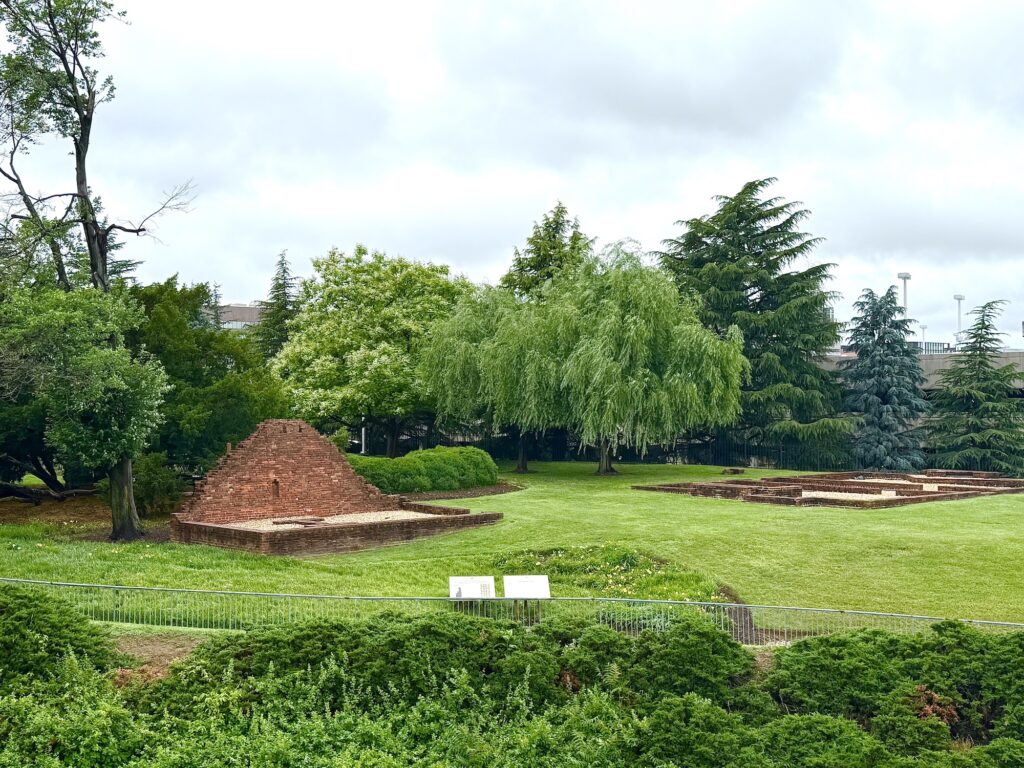 A grassy park area with brick ruins, including a small pyramid-shaped structure and rectangular foundations, surrounded by trees and shrubs under a cloudy sky.