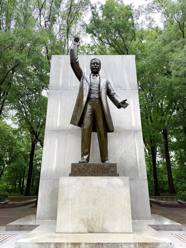 Bronze statue of Theodore Roosevelt with one arm raised, standing on a stone pedestal in front of a large stone backdrop, surrounded by trees.