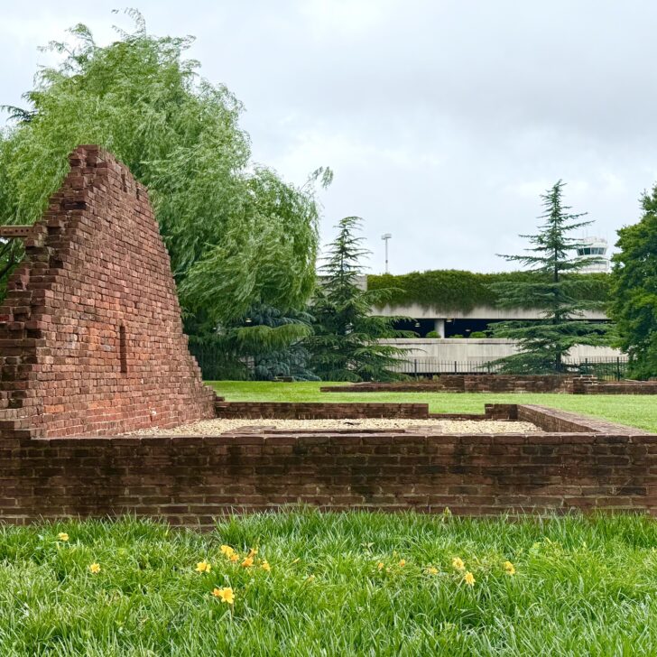 A partial brick wall from Abingdon Plantation stands alone on a grassy lawn, surrounded by trees and flowers, with an airport parking structure visible in the background.