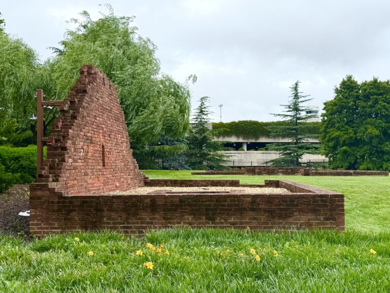 A partial brick wall from Abingdon Plantation stands alone on a grassy lawn, surrounded by trees and flowers, with an airport parking structure visible in the background.