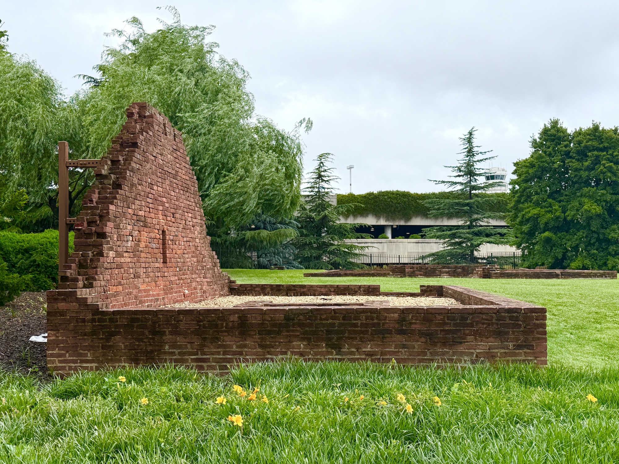 A partial brick wall from Abingdon Plantation stands alone on a grassy lawn, surrounded by trees and flowers, with an airport parking structure visible in the background.
