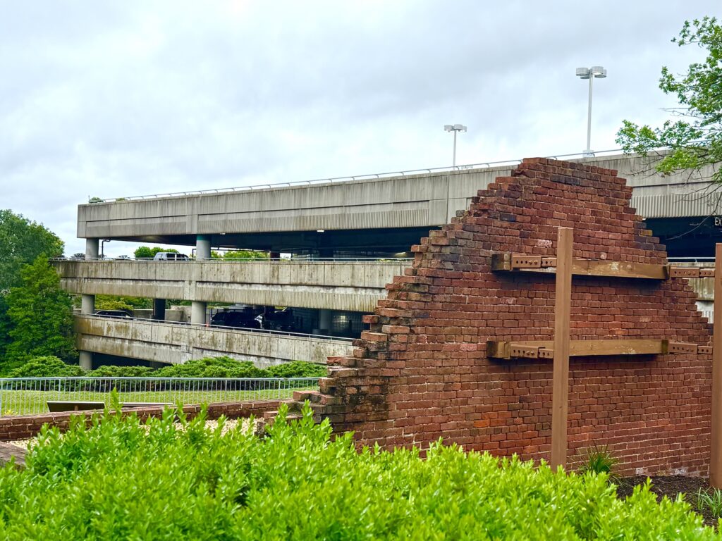 Partial red brick wall structure from the historic Abingdon Plantation stands in front of a multi-level concrete parking garage, with greenery and a cloudy sky in the background.