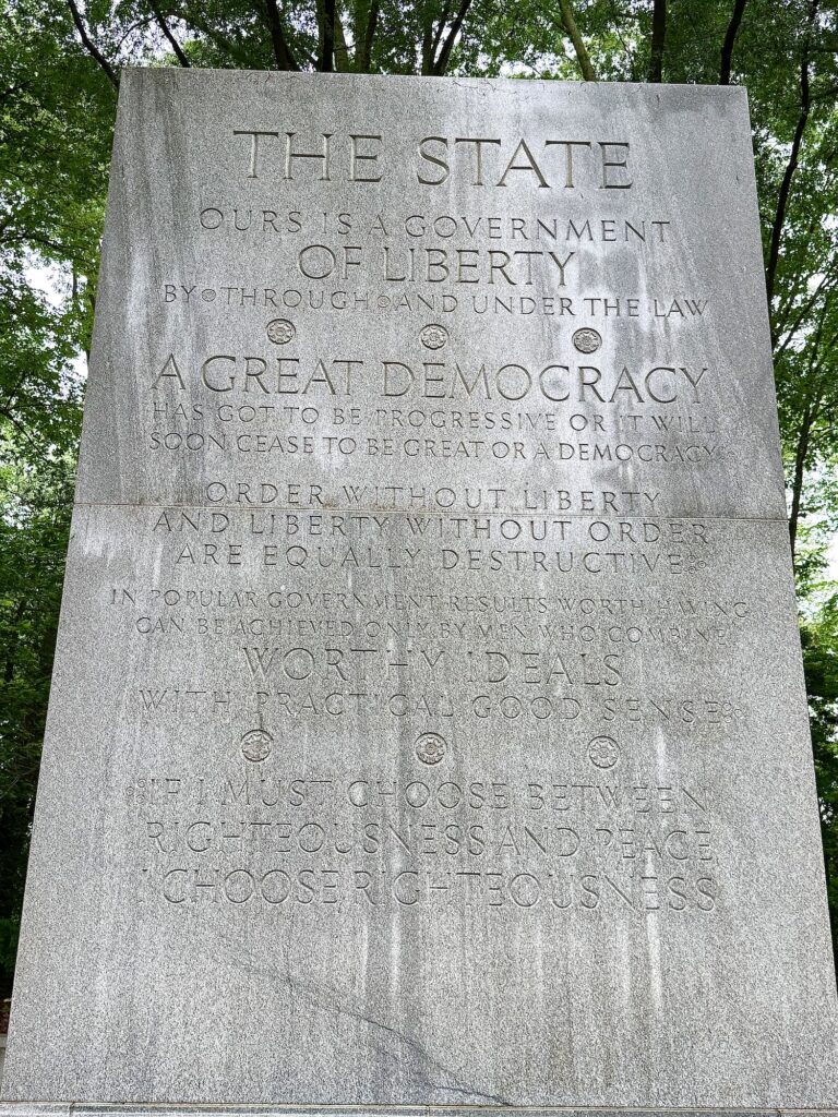 A stone monument engraved with quotes about liberty, democracy, and order, surrounded by green trees and viewed from below.