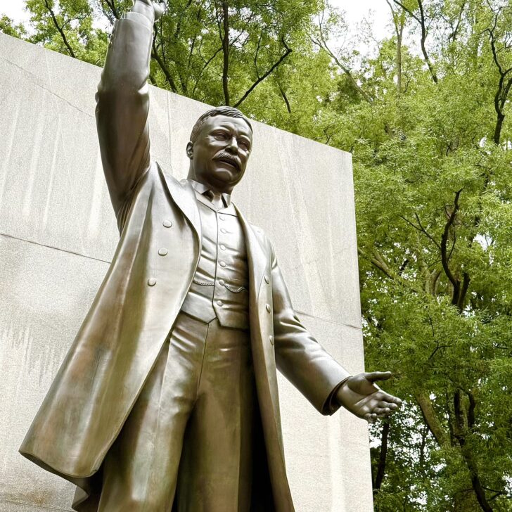 Bronze statue of President Theodore Roosevelt in a suit with one arm raised, standing in front of a stone wall and surrounded by green trees.