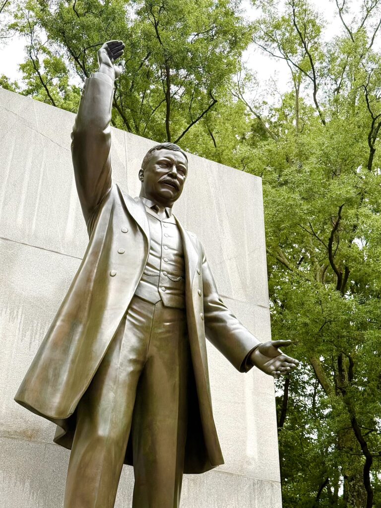Bronze statue of President Theodore Roosevelt in a suit with one arm raised, standing in front of a stone wall and surrounded by green trees.