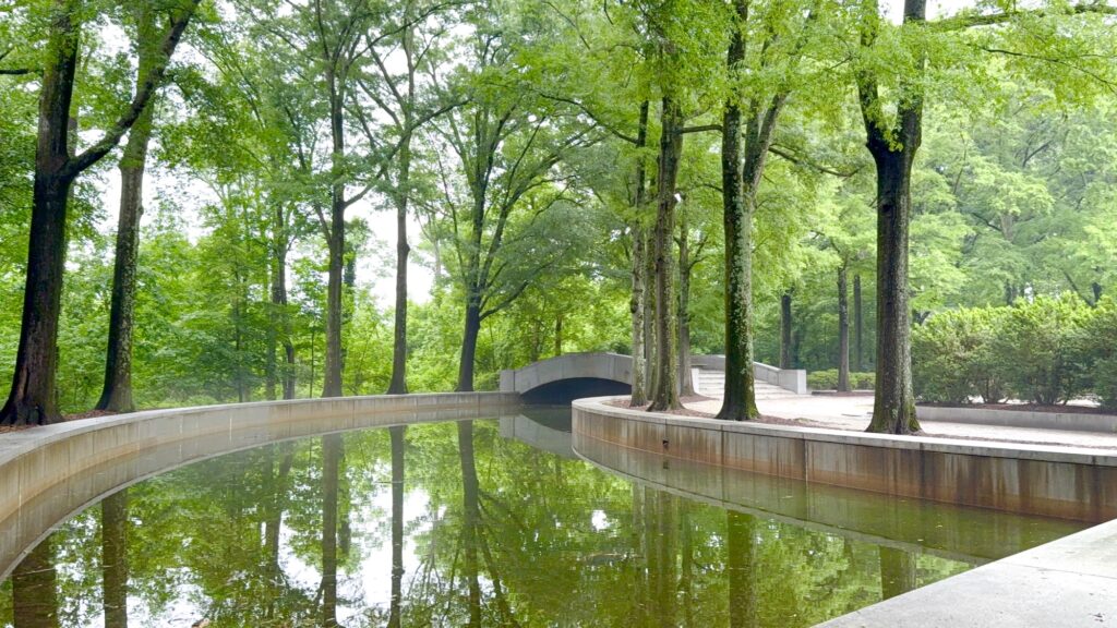 A curved canal bordered by trees reflects the greenery, with a small concrete bridge crossing over the water in a quiet park setting.