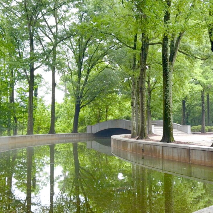 A curved canal bordered by trees reflects the greenery, with a small concrete bridge crossing over the water in a quiet park setting.
