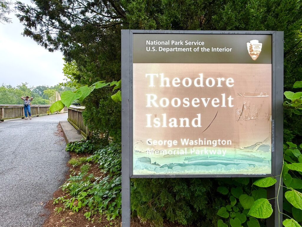 Large sign for Theodore Roosevelt Island on George Washington Memorial Parkway stands by a paved path with trees and two people in the background.