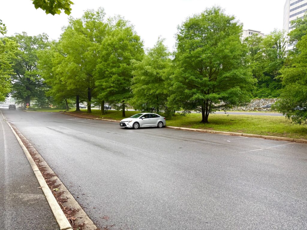 A single silver sedan is parked in an otherwise empty parking lot surrounded by green trees on a cloudy day.