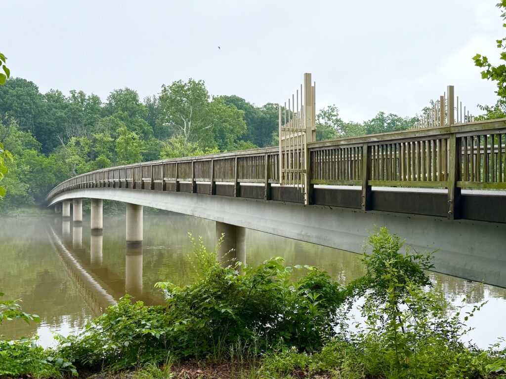 A long pedestrian bridge with metal railings spans over a calm river, surrounded by green trees and foliage on a foggy day.