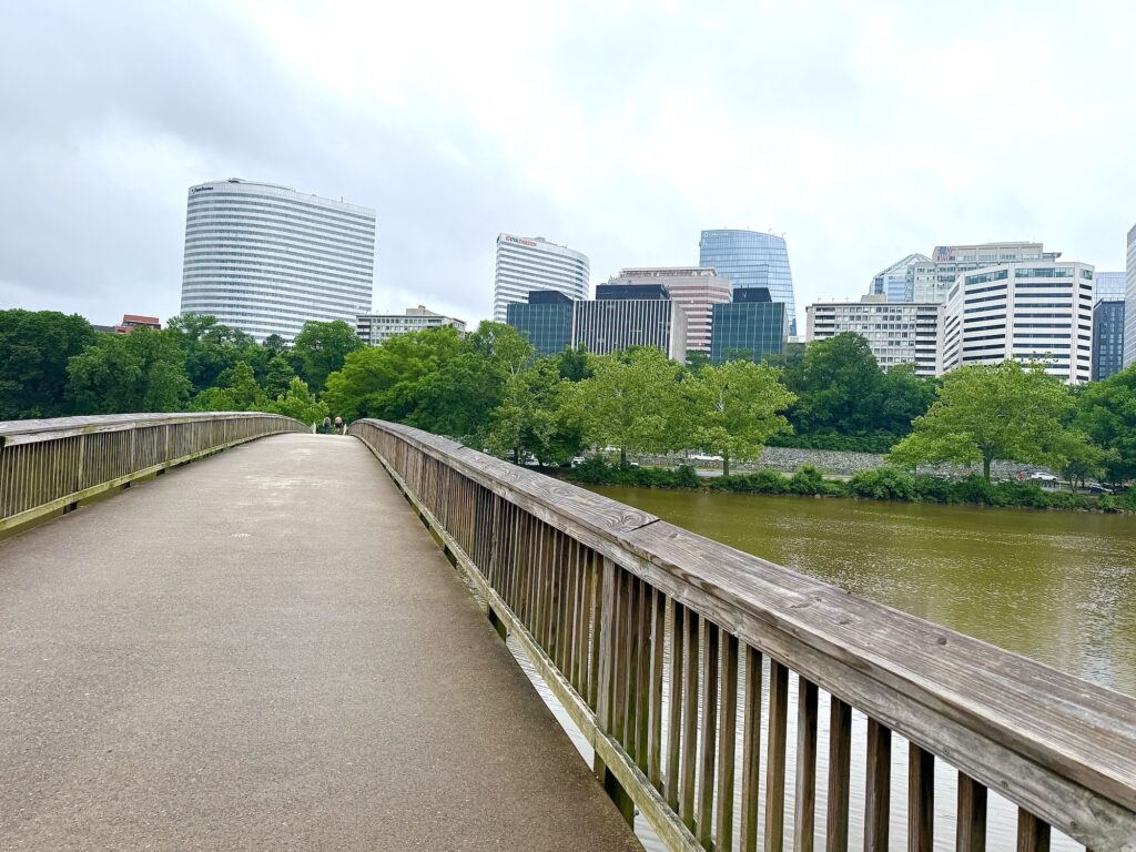 A wooden pedestrian bridge crosses over a river, with modern office buildings and green trees in the background under a cloudy sky.