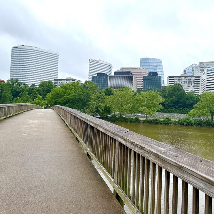 A wooden pedestrian bridge crosses over a river, with modern office buildings and green trees in the background under a cloudy sky.
