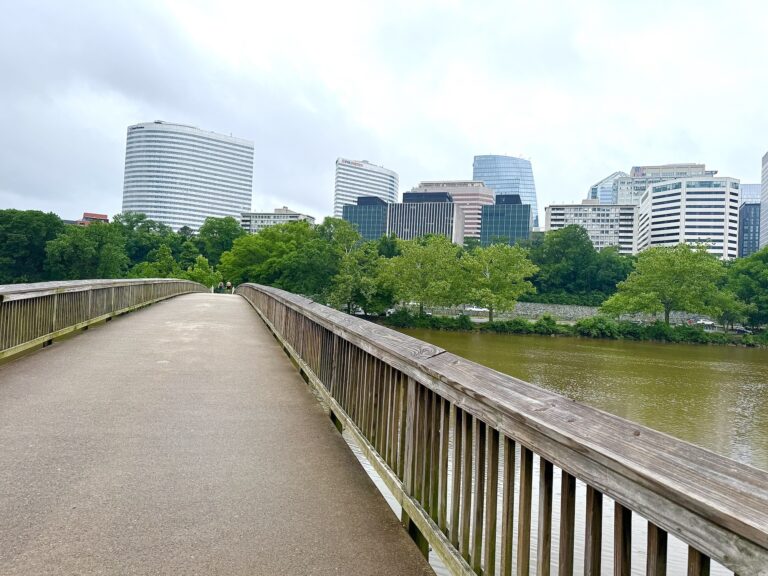 A wooden pedestrian bridge crosses over a river, with modern office buildings and green trees in the background under a cloudy sky.